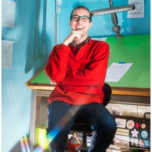 Sami Alwani wearing a red sweater and jeans. He is smiling and sitting on a chair with a drawing table behind him.