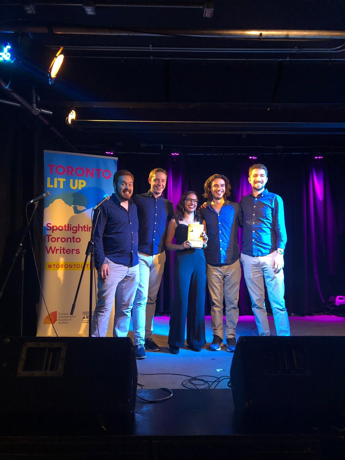 Farah Heron stands on stage with members of a barbershop quartet. Farah is holding up a copy of her book and a Toronto Lit Up banner stands behind them.