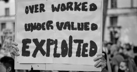 Hands are seen holding up a sign that says "Over Worked, Under Valued, Exploited"