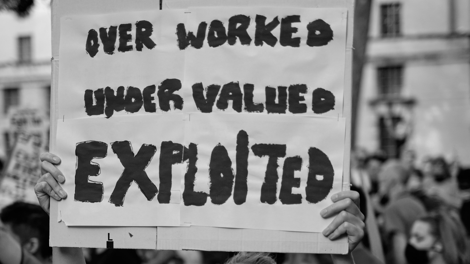 Hands are seen holding up a sign that says "Over Worked, Under Valued, Exploited"