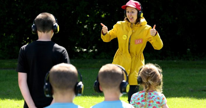 An adult in a yellow rain jacket and red baseball had is standing in front of four kids. The person is smiling with their thumbs up. Everyone is wearing black headphones outside a sunny day.