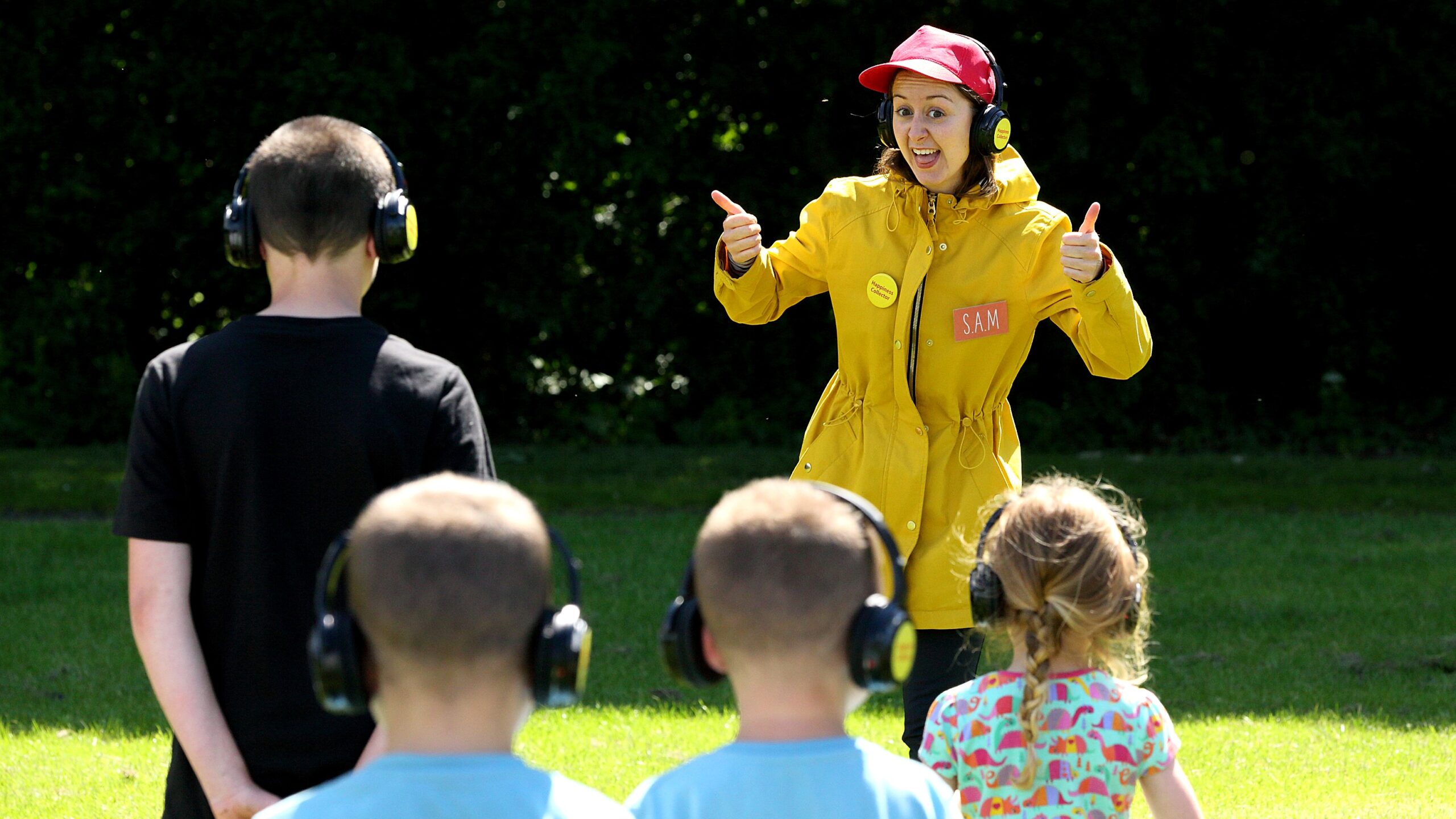 An adult in a yellow rain jacket and red baseball had is standing in front of four kids. The person is smiling with their thumbs up. Everyone is wearing black headphones outside a sunny day.