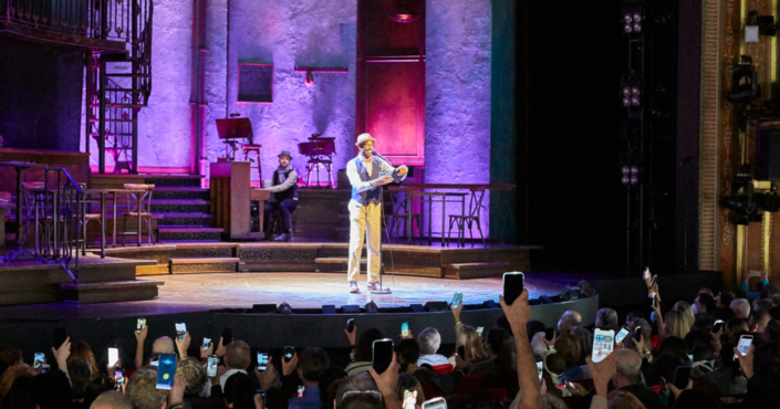 A man on stage reading with a full audience holding up their phones to shine a light on the man.