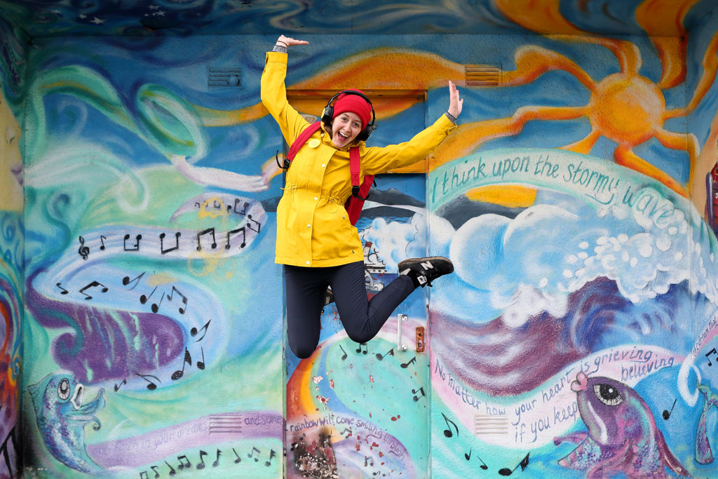 Someone in a yellow jacket jumping for joy in front of a colourful painted wall.