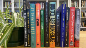 A selection of books on a table with a plant with a large bookshelf behind it.